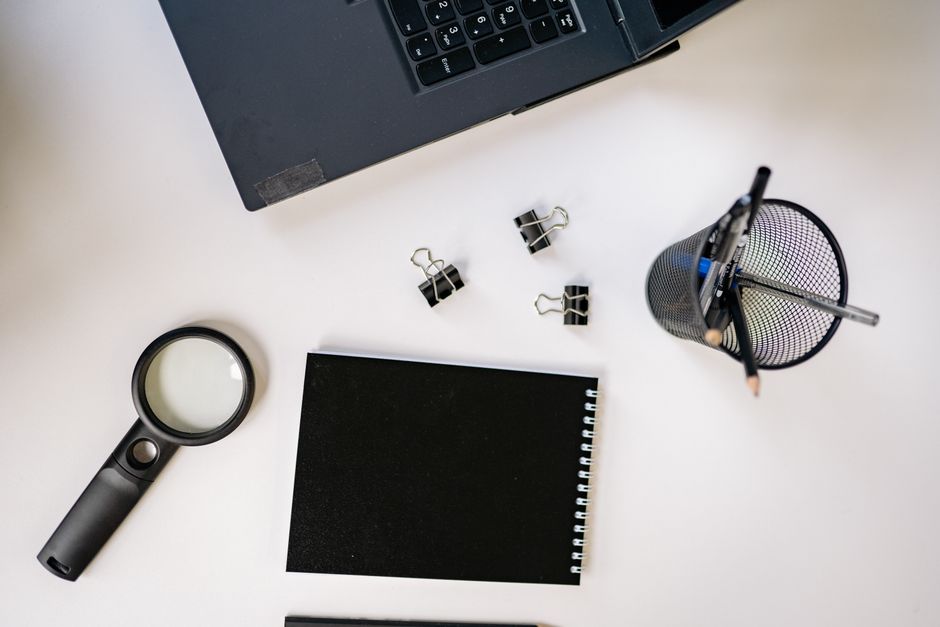 Flat lay of a workspace featuring a laptop, black notebook, magnifying glass, and office supplies on a white surface.
